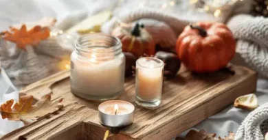 Warm autumn candle display arranged on a rustic wooden board with glowing candles, decorative pumpkins, fallen leaves, and cozy knit blankets, creating an inviting seasonal ambiance.