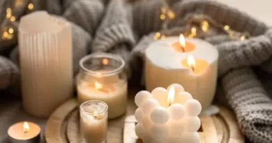 Assorted lit white/cream candles—pillar, jar, bubble-shaped, and tealight—on a wooden tray with a knit blanket and warm bokeh lights.